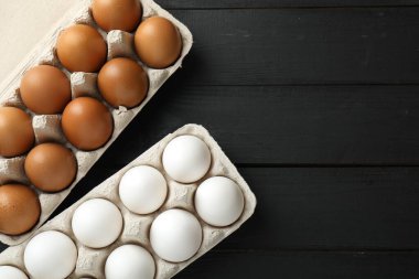 Many raw chicken eggs in cartons on black wooden table, flat lay. Space for text