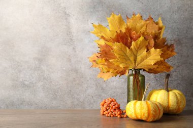 Autumn composition with vase of dry leaves, berries and pumpkins on wooden table near grey wall. Space for text