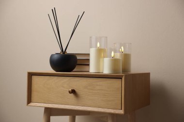 Decorative LED candles, reed diffuser and books on wooden bedside table near beige wall indoors