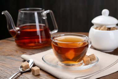 Aromatic black tea in glass cup, brown sugar and teapot on wooden table, closeup