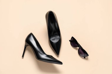 Bir çift zarif siyah topuklu ve bej arka planda güneş gözlüğü, düz bir şekilde yatıyordu. Şık ayakkabılar.