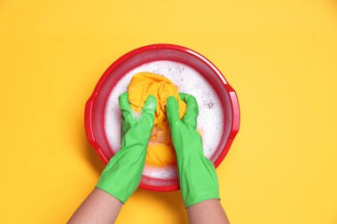 Sarı arka planda plastik leğende çamaşırları yıkayan kadın.