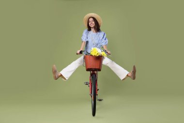 Yeşil arka planda bir sepet çiçekle bisiklete binen şapkalı gülümseyen kadın.
