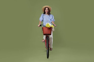 Yeşil arka planda bir sepet çiçekle bisiklete binen şapkalı gülümseyen kadın.