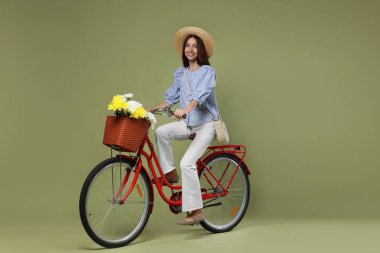Yeşil arka planda bir sepet çiçekle bisiklete binen şapkalı gülümseyen kadın.