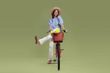 Yeşil arka planda çiçek sepeti olan şapkalı ve bisikletli gülümseyen kadın.