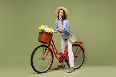 Yeşil arka planda çiçek sepeti olan şapkalı ve bisikletli gülümseyen kadın.