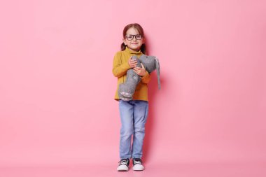 Gözlüklü, pembe arka planda oyuncak tavşanı olan sevimli bir kız.