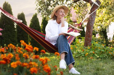 Hasır şapkalı ve kitaplı yaşlı kadın dışarıda hamakta dinleniyor.