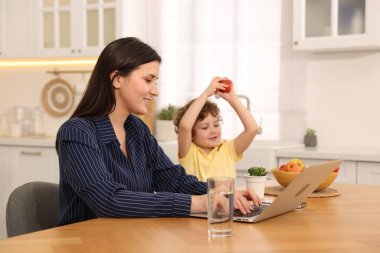 Oğlu ev ofisinin ahşap masasında elmayla oynarken dizüstü bilgisayarla çalışan gülümseyen kadın.