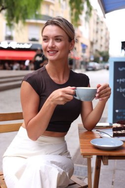 Dışarıdaki ahşap masada bir fincan kahve ve bir dilim pastayla güzel genç bir kadın.