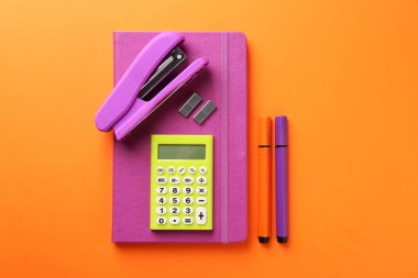 Stapler, metal staples, notebook, calculator and felt pens on orange background, flat lay