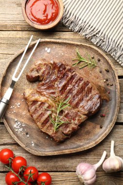 Delicious grilled beef steak with rosemary, spices, ketchup, carving fork and products on wooden table, flat lay