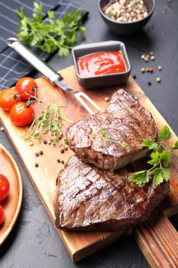 Delicious grilled beef steaks with herbs, peppercorns, tomatoes, carving fork and ketchup on dark textured table, closeup