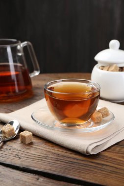 Aromatic black tea in glass cup, brown sugar and teapot on wooden table, closeup
