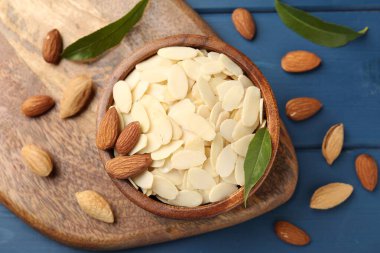 Fresh almond flakes, whole nuts and green leaves on blue wooden table, flat lay