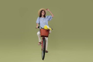 Yeşil arka planda bir sepet çiçekle bisiklete binen şapkalı gülümseyen kadın.