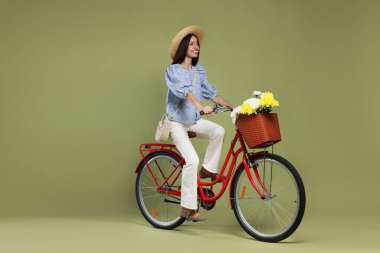 Yeşil arka planda bir sepet çiçekle bisiklete binen şapkalı gülümseyen kadın. Metin için boşluk