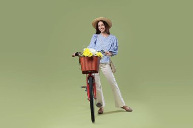 Yeşil arka planda çiçek sepeti olan şapkalı ve bisikletli gülümseyen kadın.