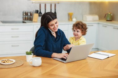 Akıllı telefonuyla konuşan gülümseyen bir kadın dizüstü bilgisayarla çalışırken oğlu da ofis odasındaki ahşap masada.