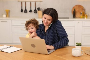 Akıllı telefonuyla konuşan gülümseyen bir kadın dizüstü bilgisayarla çalışırken oğlu da ofis odasındaki ahşap masada.