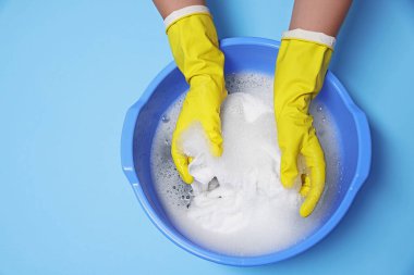 Elbiseleri açık mavi arka planda plastik bir leğende yıkayan kadın.