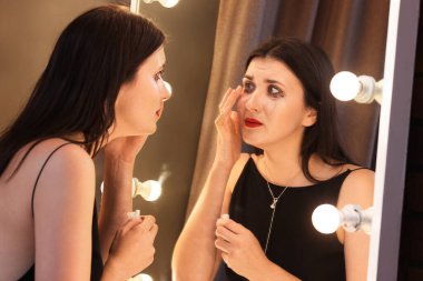 Elbisesine maskara bulaşmış bir kadın aynanın yanında gözyaşlarını siliyor.