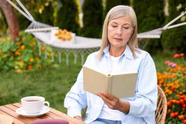 Bahçedeki ahşap masada kitap okuyan yaşlı bir kadın.