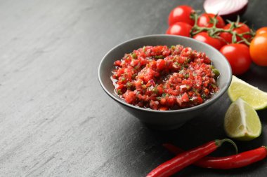 Delicious salsa sauce in bowl and ingredients on dark grey table, closeup. Space for text