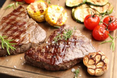 Delicious grilled beef steaks with herbs and vegetables on wooden board, closeup