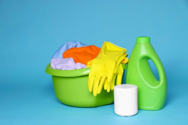 Açık mavi arka planda çamaşır, deterjan ve lastik eldivenli plastik bir leğen.