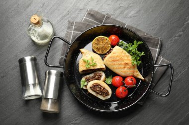 Pieces of tasty grilled squid stuffed with mushrooms, parsley, tomatoes and lemon in pan on dark textured table, flat lay