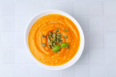 Delicious pumpkin soup with seeds and basil in bowl on white tiled table, top view
