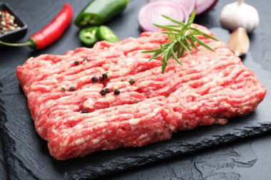 Raw minced meat with rosemary, spices, onion and chili peppers on black textured table, closeup