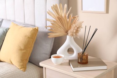 Evde yatağın yanında bir fincan çay, kitap ve dekor olan komodin.