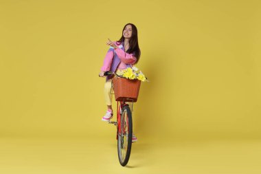 Bir sepet çiçekle bisiklete binen gülümseyen kadın ve sarımsı yeşil arka planda bir şeye işaret eden.