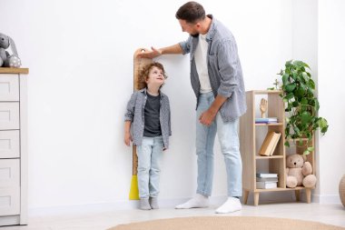 Baba, evdeki beyaz duvarın yanında küçük oğlunun boyunu ölçüyor.