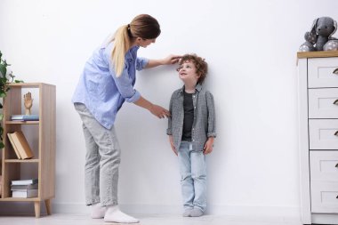 Anne, küçük oğlunun boyunu içeride beyaz duvarın yanında ölçüyor.