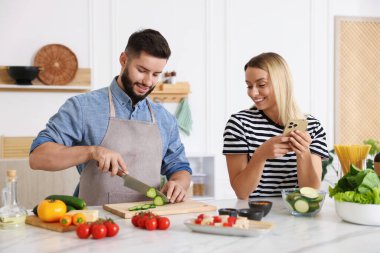 Yemek pişirme süreci. Karısı mutfaktaki masada akıllı telefondan yemek tarifine bakarken gülümseyen adam salatalık kesiyor.
