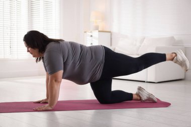 Evde yoga minderinde esneyen spor kıyafetli şişman bir kadın.