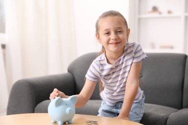 Küçük kız cep harçlığını ahşap masadaki domuz kumbarasına yatırıyor.