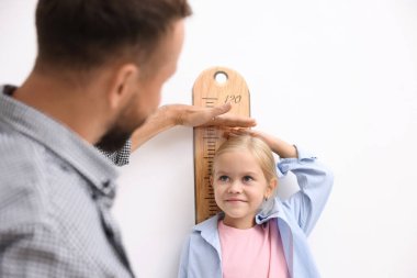 Babası küçük kızının boyunu ölçüyor. Beyaz duvarın yanında.