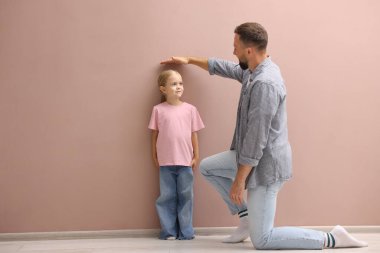 Babası küçük kızının boyunu ölçüyor. Beyaz bir duvarın yanında..