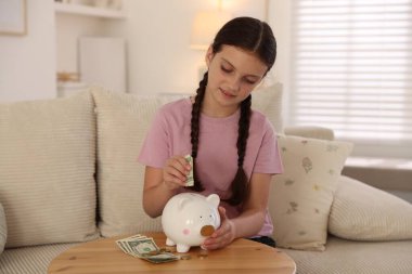 Küçük kız evdeki kanepedeki ahşap masadaki domuz kumbarasına para yatırıyor. Cep harçlığı ve sorumluluk