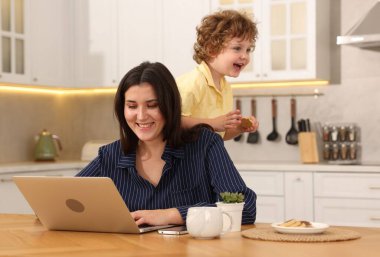 Oğlu ev ofisinin ahşap masasında kurabiyeyle oynarken dizüstü bilgisayarla çalışan gülümseyen kadın.