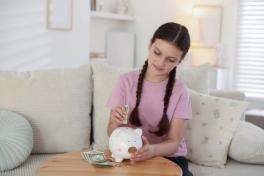Küçük kız evdeki kanepedeki ahşap masadaki domuz kumbarasına para yatırıyor. Cep harçlığı ve sorumluluk