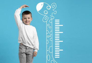 Küçük çocuk boyunu açık mavi arkaplanda ölçüp zürafa çiziyor.