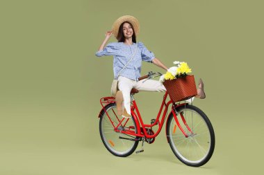 Yeşil arka planda bir sepet çiçekle bisiklete binen şapkalı gülümseyen kadın. Metin için boşluk