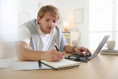 İçerideki ahşap masada dizüstü bilgisayar kullanırken not alan adam.