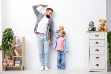 Baba ve kızı evdeki beyaz duvarın yanında boylarını ölçüyorlar.